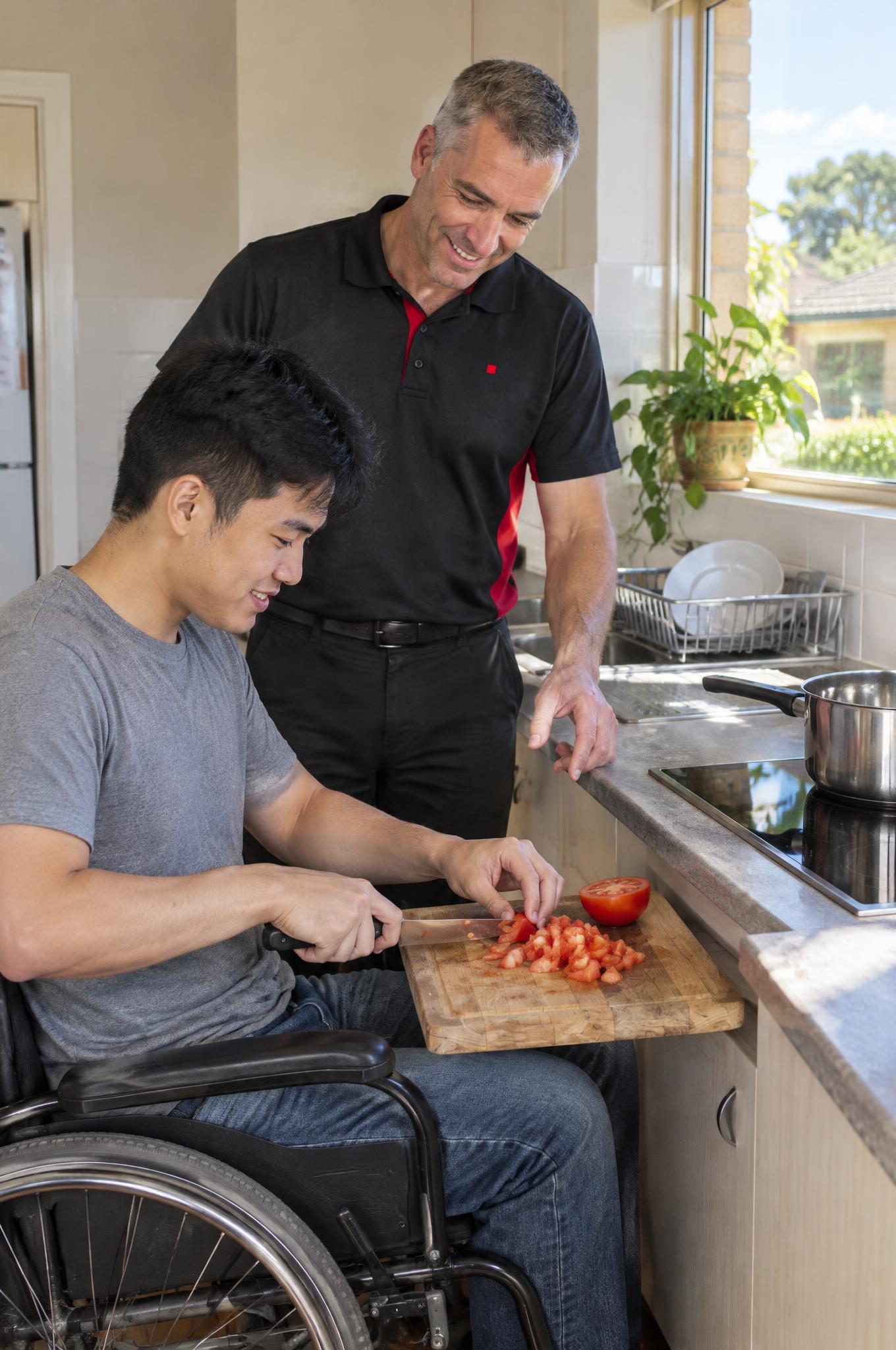 Young man in a wheelchair chopping tomatoes at his kitchen bench while an occupational therapist looks on