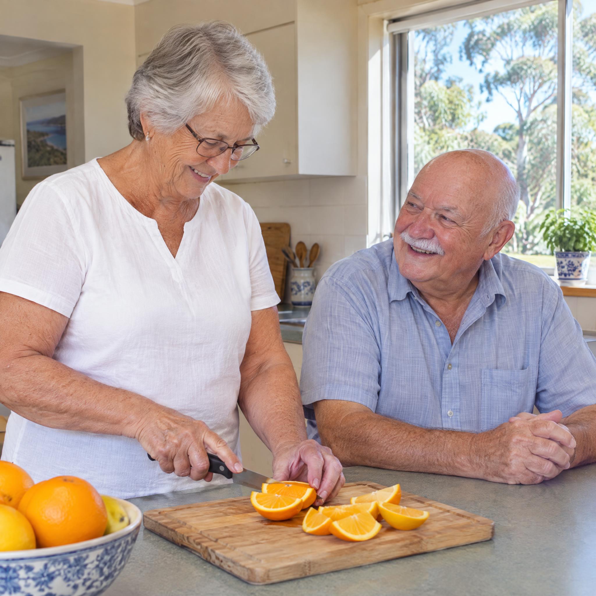 Older woman slicing oranges at her kitchen bench while her husband chats beside her