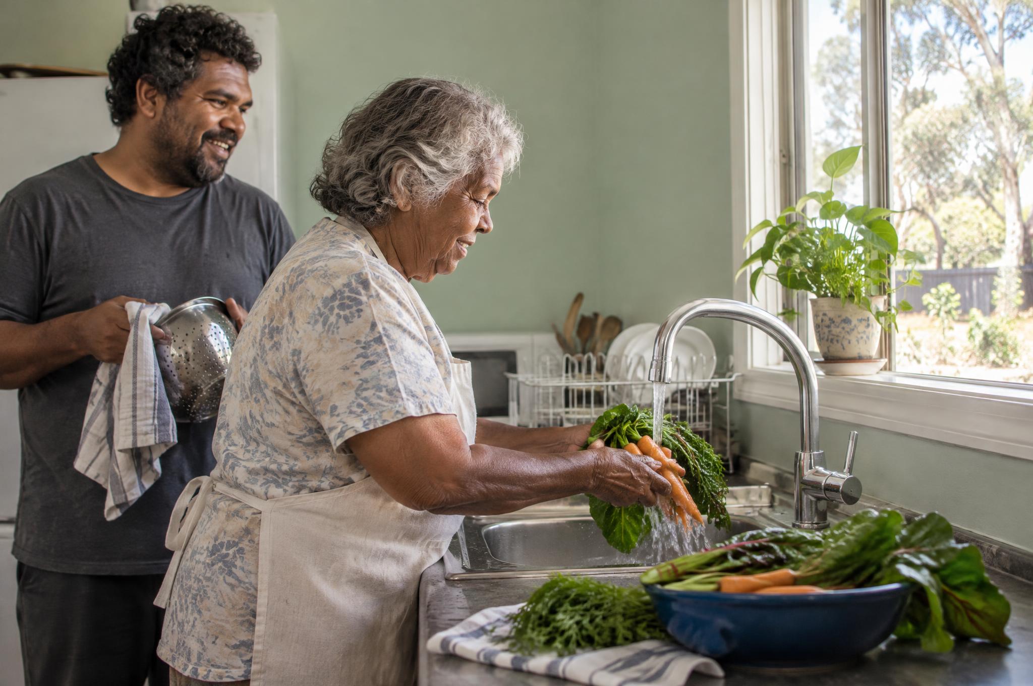 Older woman washing carrots and greens at her kitchen sink while her adult son dries a colander beside her
