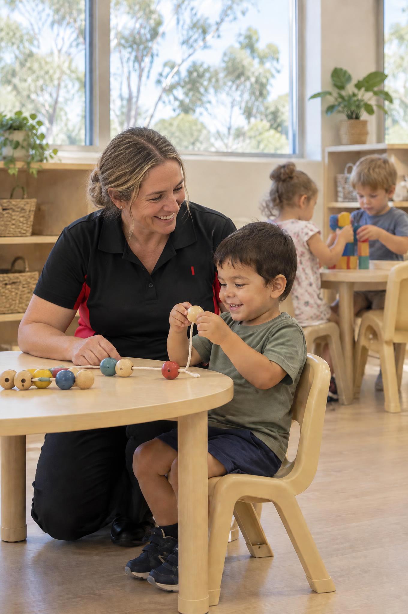 Paediatric occupational therapist kneeling beside a young boy threading wooden beads at a kindy table
