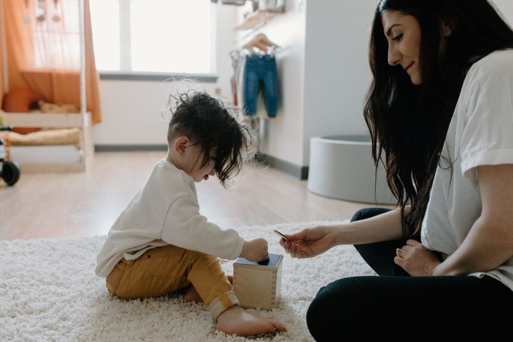 Mother sitting on a rug beside her toddler, guiding him to post a coin into a wooden box during play-based fine motor activity at home