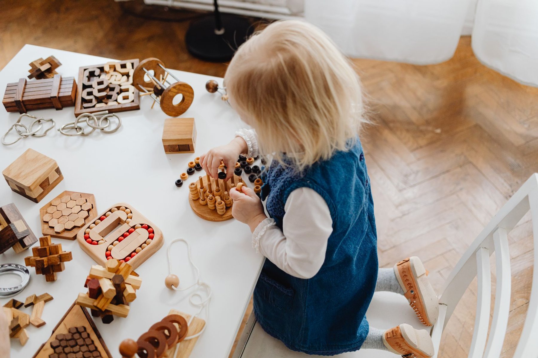 Toddler kneeling at a white table filled with wooden fine motor toys, placing beads on pegs in a bright home play space