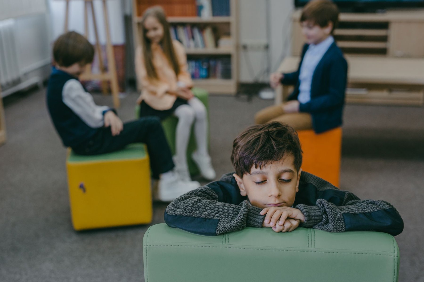 Quiet boy resting his chin on a chair in a classroom while other children talk in the background, showing the social withdrawal linked to selective mutism