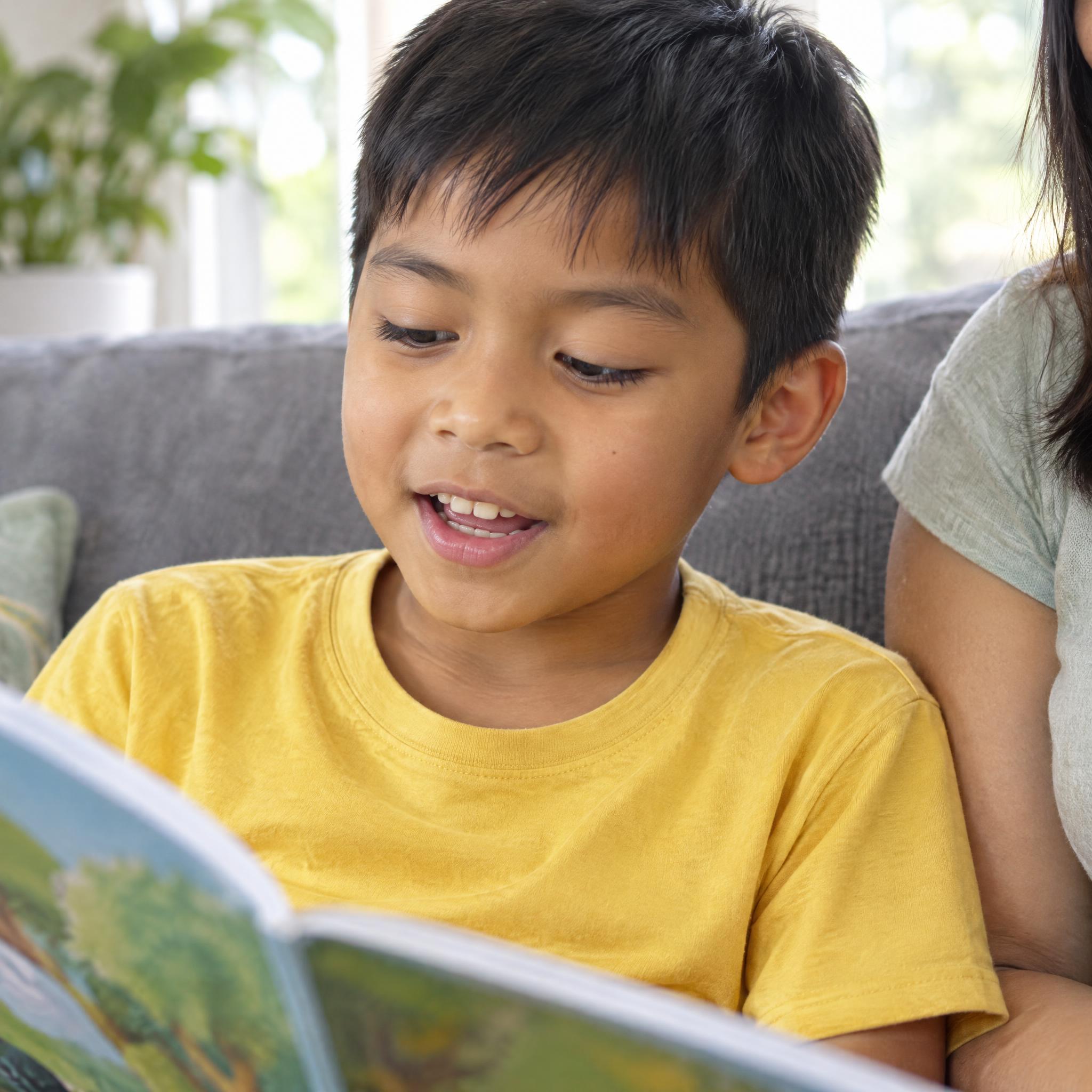 Young boy reading a picture book aloud on the couch at home with his mum beside him